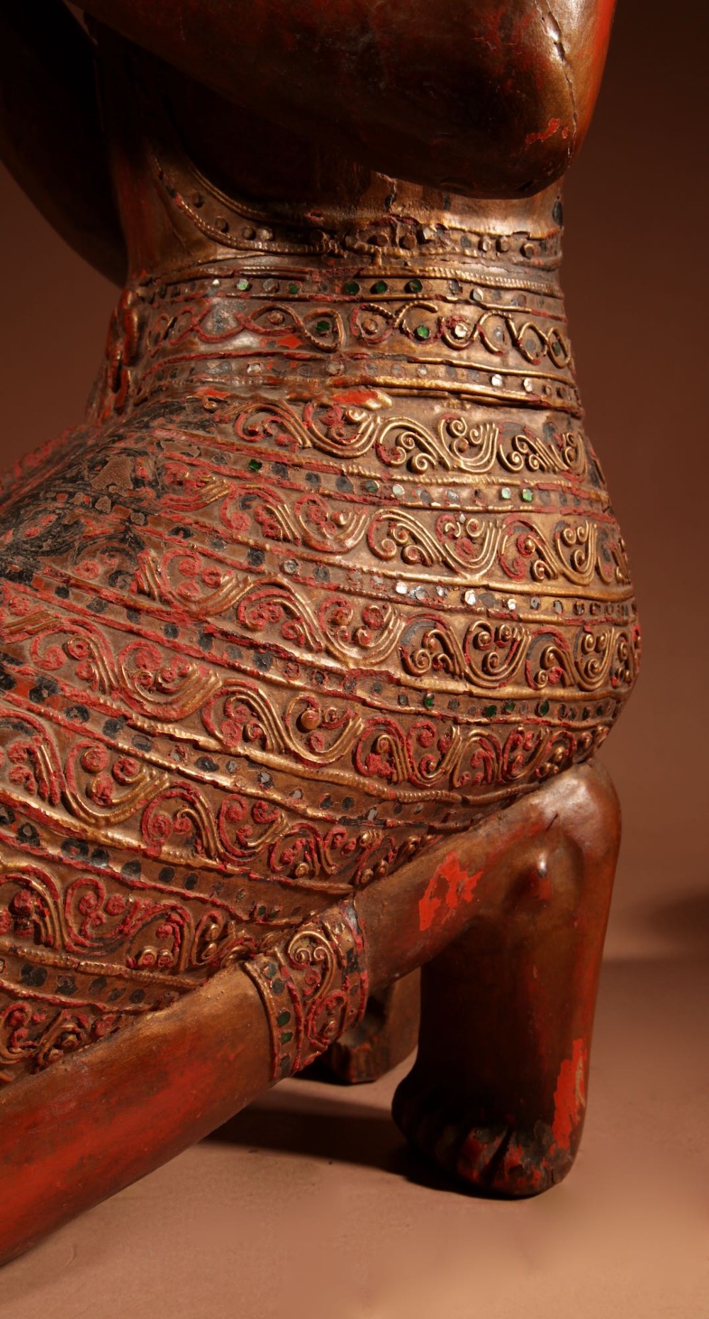 A carved wooden Thai Thepphanom guardian statue depicted kneeling with hands held in a prayer gesture, featuring a conical headdress and gilded red lacquered details. View IX