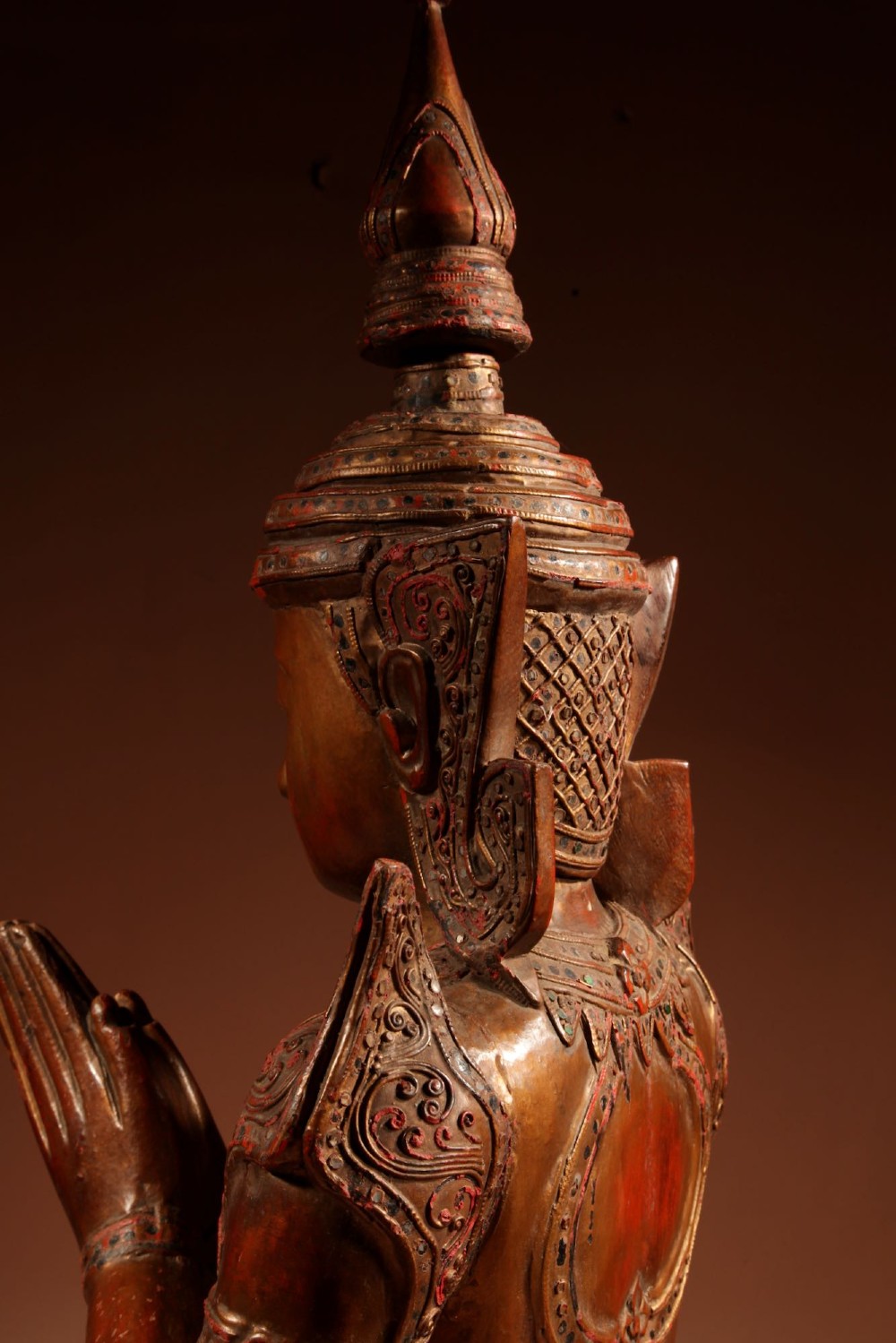 A carved wooden Thai Thepphanom guardian statue depicted kneeling with hands held in a prayer gesture, featuring a conical headdress and gilded red lacquered details. View X