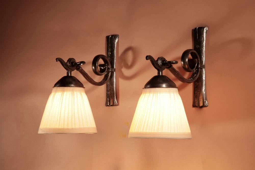 A pair of Art Deco wrought iron wall sconces featuring curved scroll arms and conical pleated cream fabric shades mounted on wall brackets.