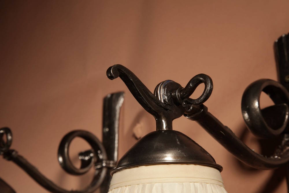 A pair of Art Deco wrought iron wall sconces featuring curved scroll arms and conical pleated cream fabric shades mounted on wall brackets. View VI