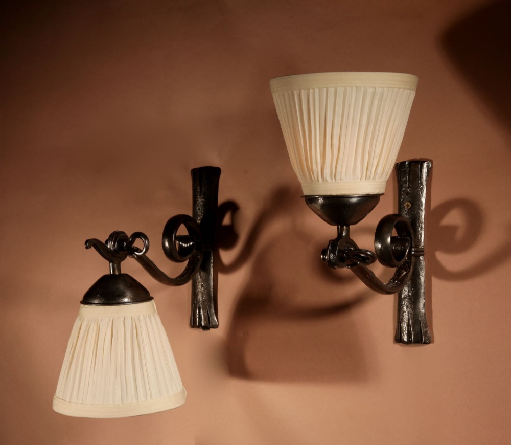 A pair of Art Deco wrought iron wall sconces featuring curved scroll arms and conical pleated cream fabric shades mounted on wall brackets. View IX