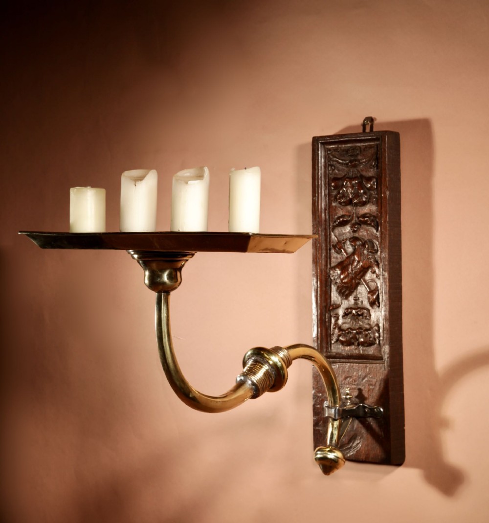 A wall-mounted candle sconce featuring a carved dark oak backboard and a curved brass arm holding a tray with four white candles.