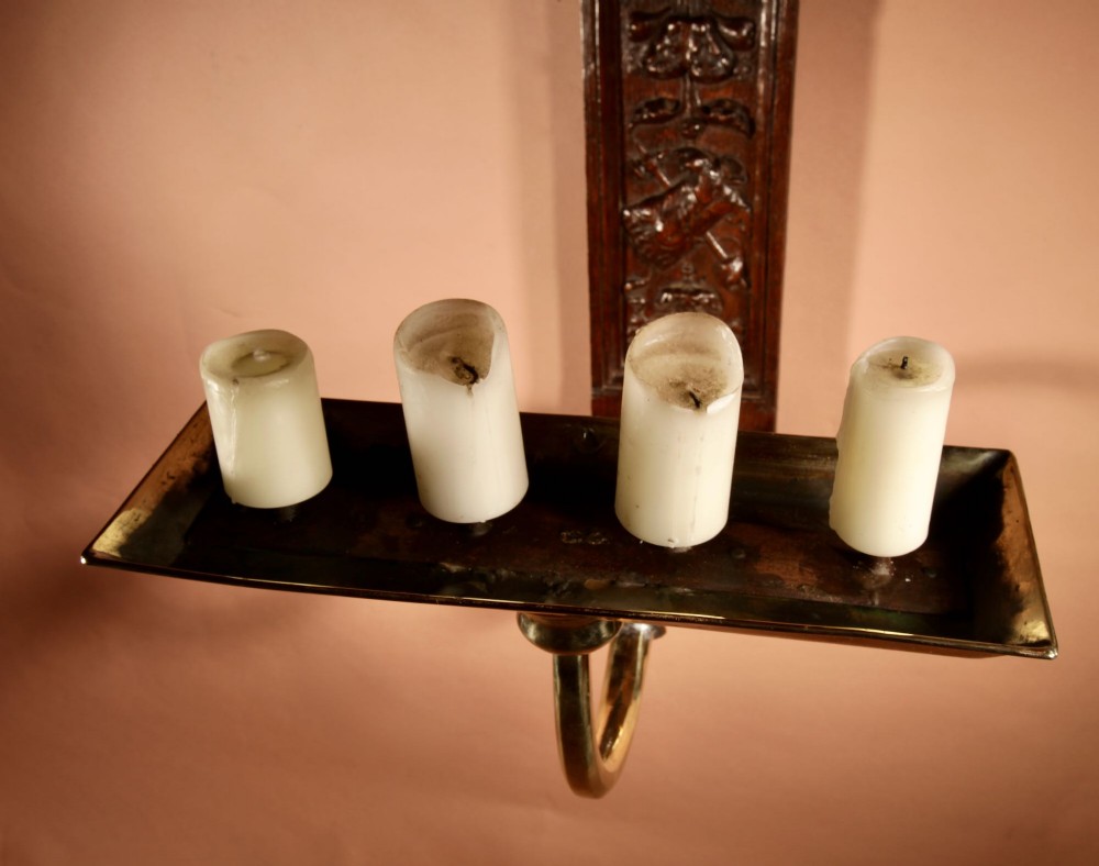 A wall-mounted candle sconce featuring a carved dark oak backboard and a curved brass arm holding a tray with four white candles. View X