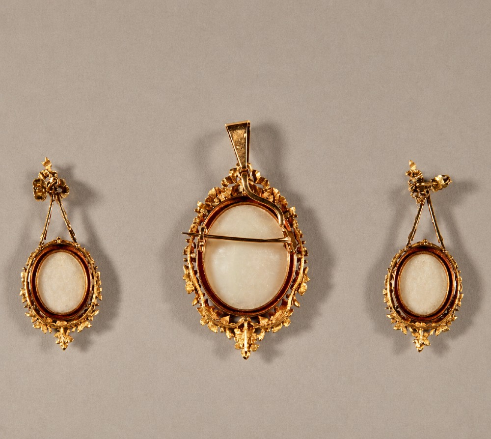 A three-piece antique gold demi-parure comprising an oval pendant and matching drop earrings featuring hand-painted porcelain miniatures of cherubs and classical figures framed by intricate leafy gold scrollwork. View II
