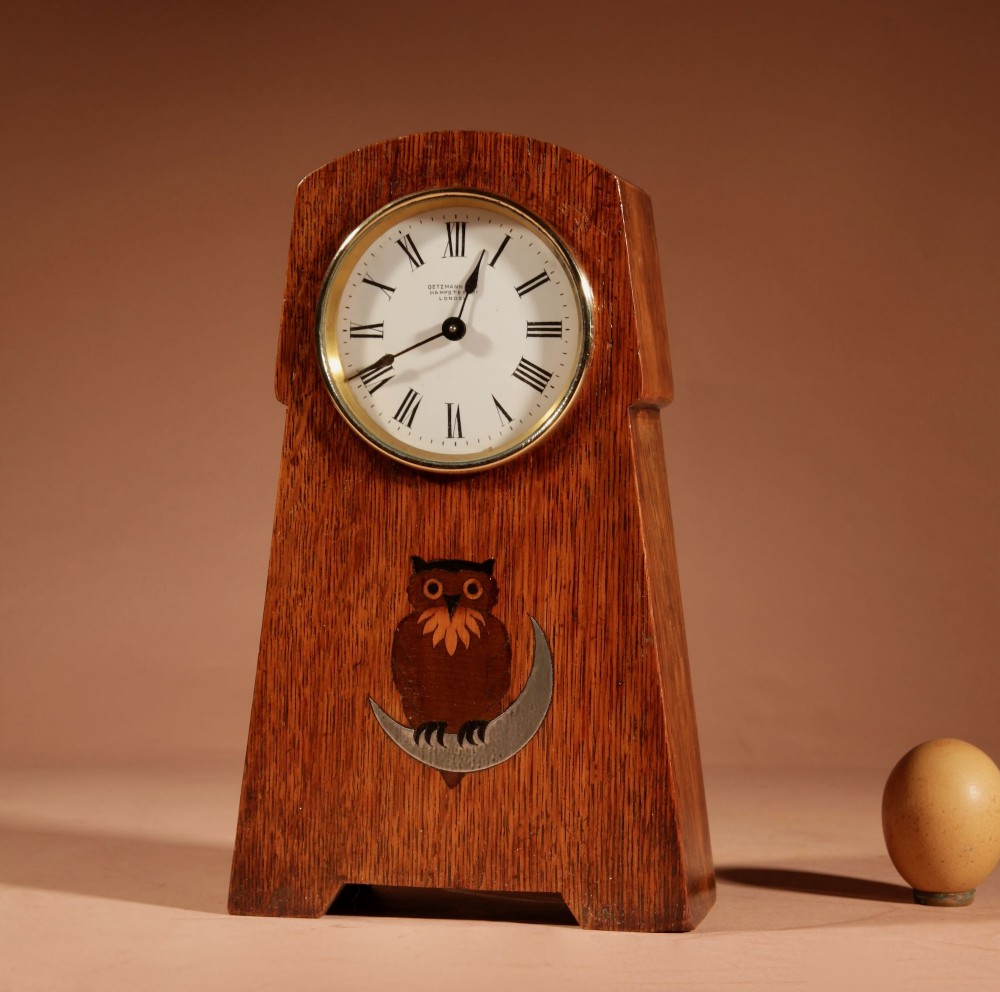 An Arts and Crafts tapered oak mantel clock featuring a white circular dial and wood inlay depicting an owl perched on a pewter crescent moon.