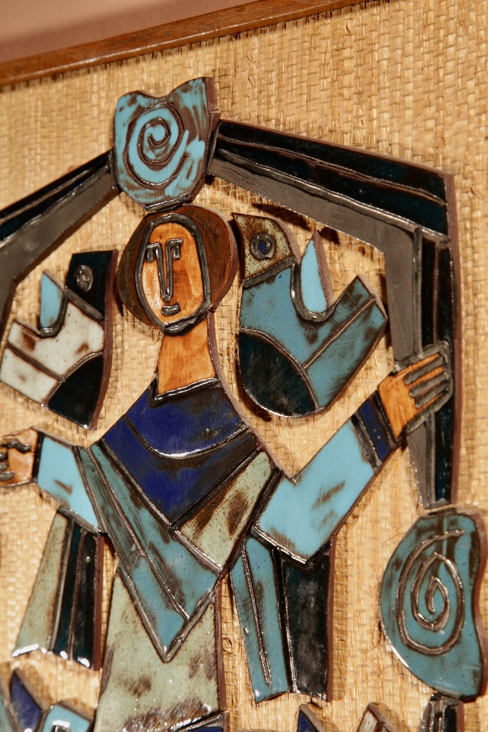 A framed mid-century modern ceramic wall relief featuring stylised figures and birds in turquoise and blue glazes mounted on a woven background. View V