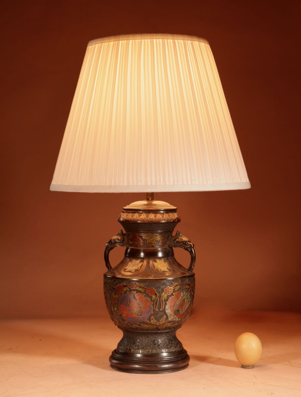 A Japanese champleve bronze urn formatted as a table lamp with polychrome enamel floral patterns, sculpted handles, and a cream pleated shade.