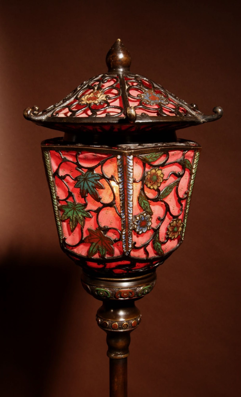 A tall Japanese bronze standard lamp featuring an illuminated red glass lantern shade with enamelled foliage motifs supported on a slender column base. View II