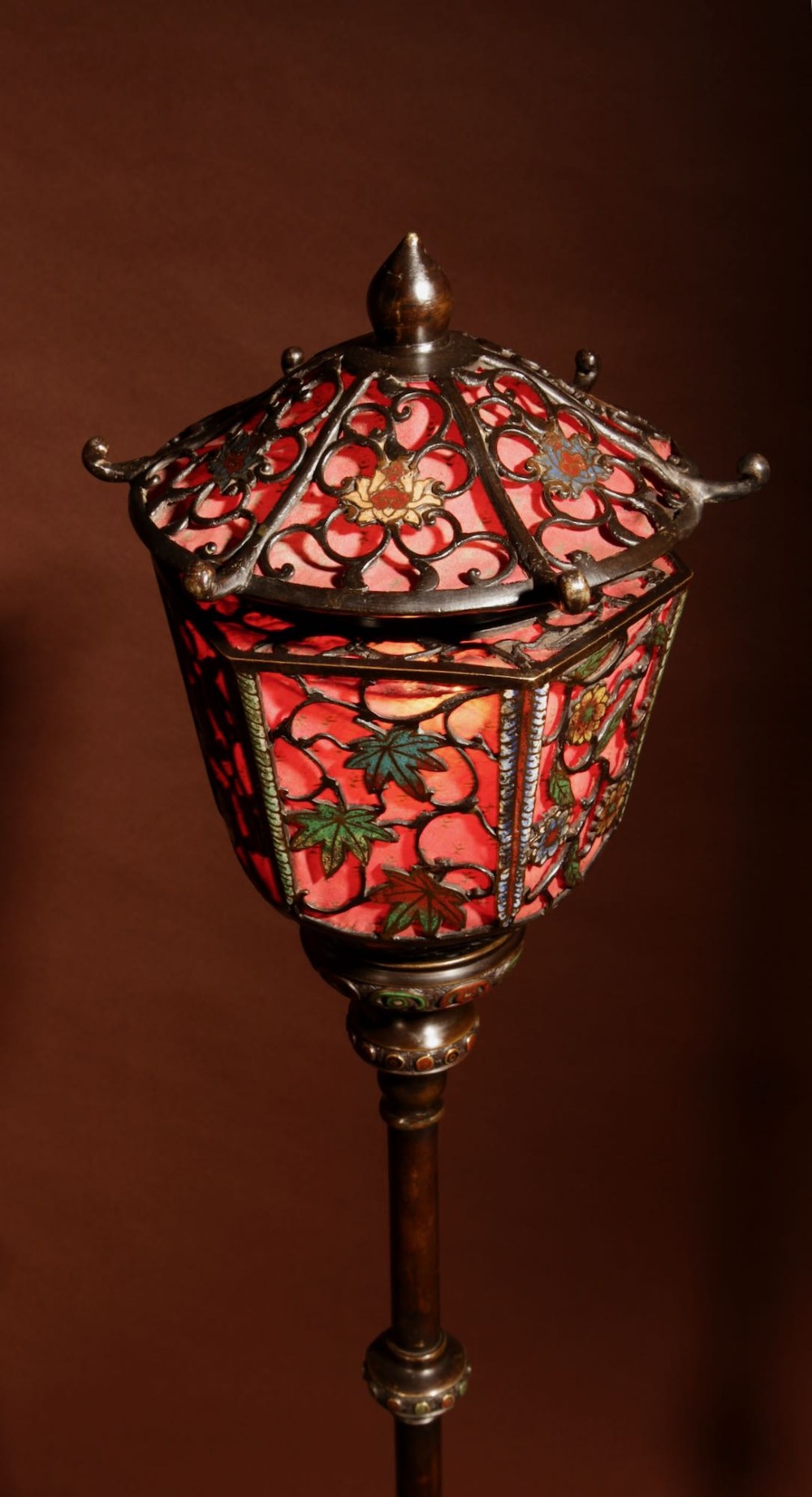 A tall Japanese bronze standard lamp featuring an illuminated red glass lantern shade with enamelled foliage motifs supported on a slender column base. View III