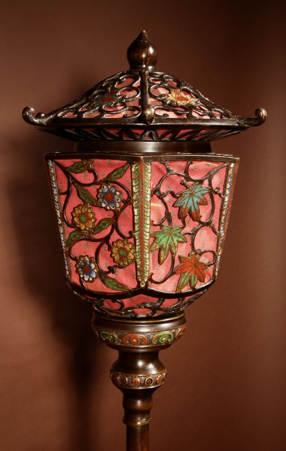 A tall Japanese bronze standard lamp featuring an illuminated red glass lantern shade with enamelled foliage motifs supported on a slender column base. View VI