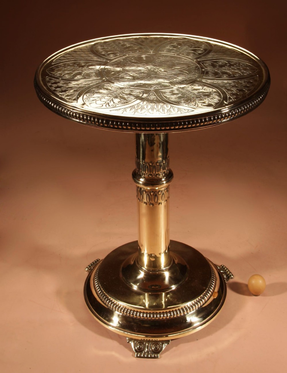 A circular polished brass side table featuring an embossed Art Nouveau floral pattern on top, supported by a central column pedestal and rounded base.