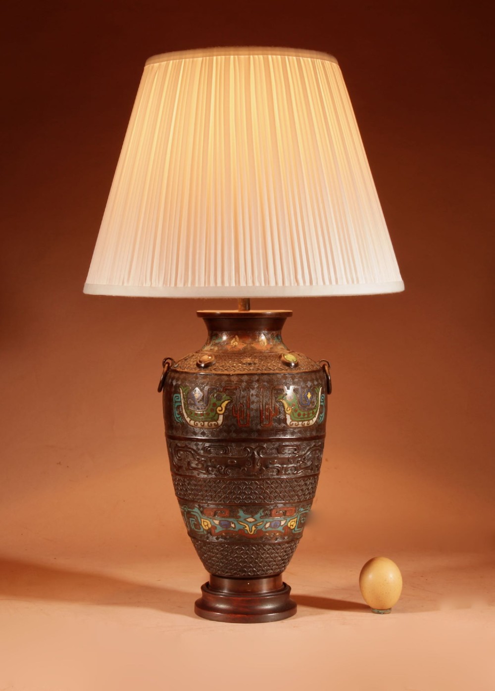 A Japanese cast bronze table lamp decorated with polychrome champleve enamel band motifs and fitted with a pleated cream shade.