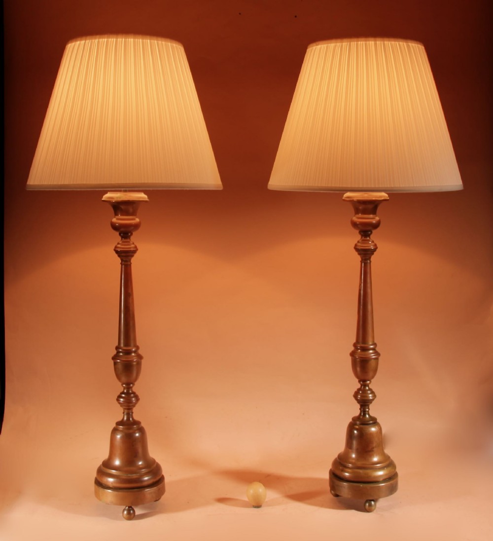 A pair of tall patinated brass candlestick table lamps featuring turned stems and pleated cream shades on circular bases.