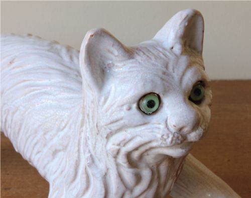fig 1. A cream-glazed terracotta cat figure depicted in a crouching pose featuring bright green glass eyes.