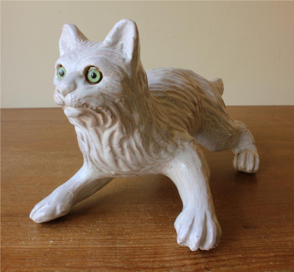 A cream-glazed terracotta cat figure depicted in a crouching pose featuring bright green glass eyes.