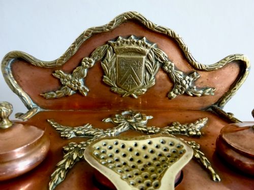 fig 1. An antique copper and brass inkstand featuring a contoured backplate with a heraldic crest, brass swags, two capped inkwells, and brass hoof feet.
