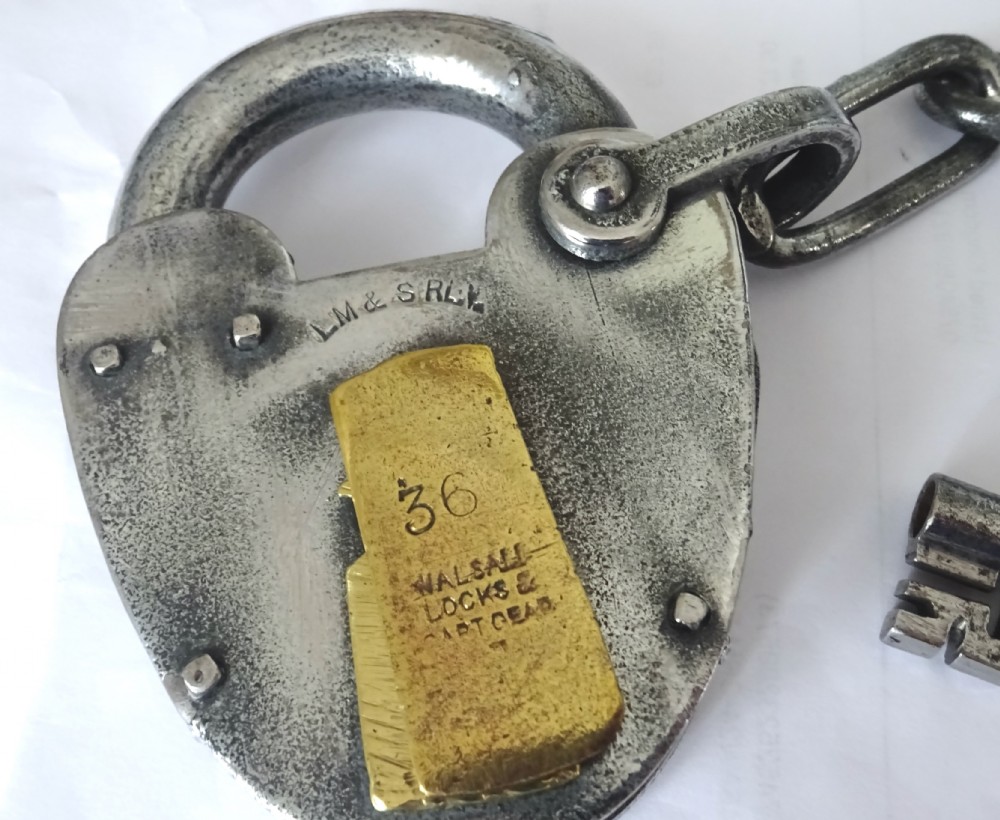 An antique heart-shaped steel railway padlock featuring a stamped brass keyhole cover, attached chain, and matching iron key. View V
