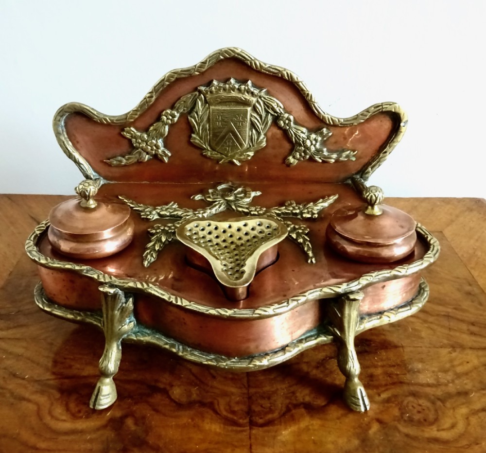 An antique copper and brass inkstand featuring a contoured backplate with a heraldic crest, brass swags, two capped inkwells, and brass hoof feet.
