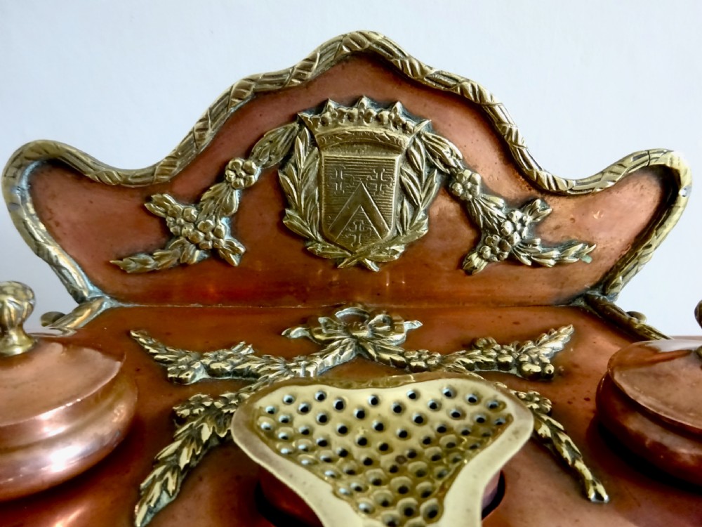 An antique copper and brass inkstand featuring a contoured backplate with a heraldic crest, brass swags, two capped inkwells, and brass hoof feet. View II