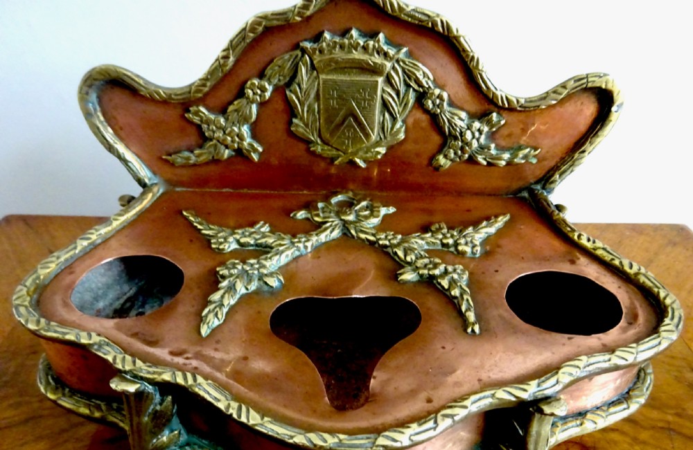 An antique copper and brass inkstand featuring a contoured backplate with a heraldic crest, brass swags, two capped inkwells, and brass hoof feet. View III