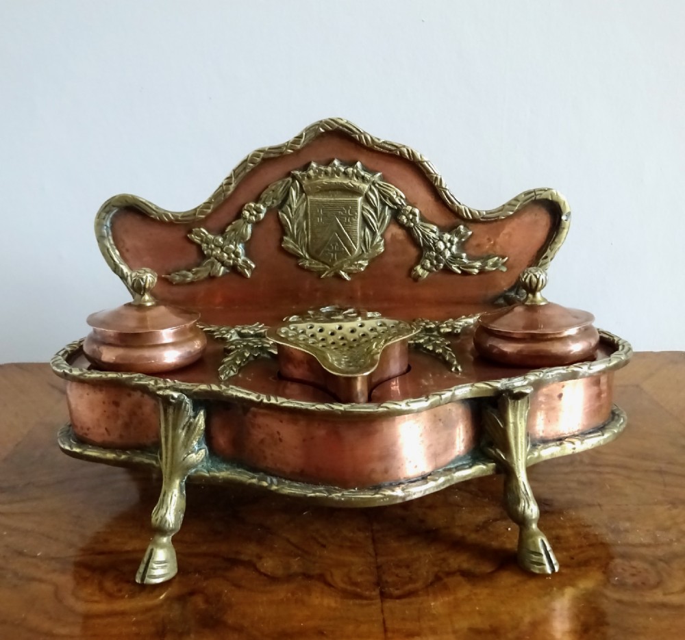 An antique copper and brass inkstand featuring a contoured backplate with a heraldic crest, brass swags, two capped inkwells, and brass hoof feet. View IV