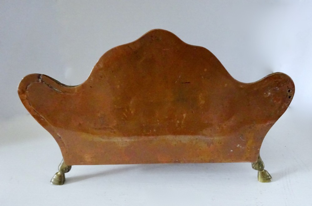 An antique copper and brass inkstand featuring a contoured backplate with a heraldic crest, brass swags, two capped inkwells, and brass hoof feet. View V