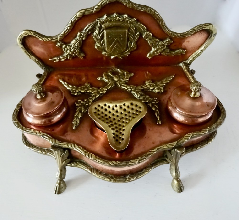 An antique copper and brass inkstand featuring a contoured backplate with a heraldic crest, brass swags, two capped inkwells, and brass hoof feet. View VI
