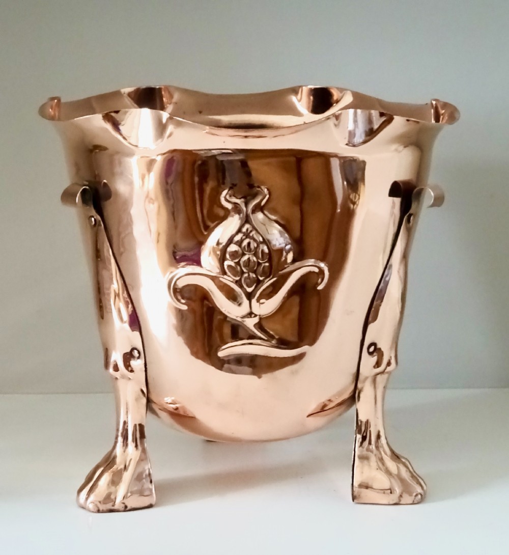 A polished copper Arts and Crafts jardiniere with a scalloped frilled rim, repousse botanical seed pod motif, and riveted metal legs terminating in paw feet.