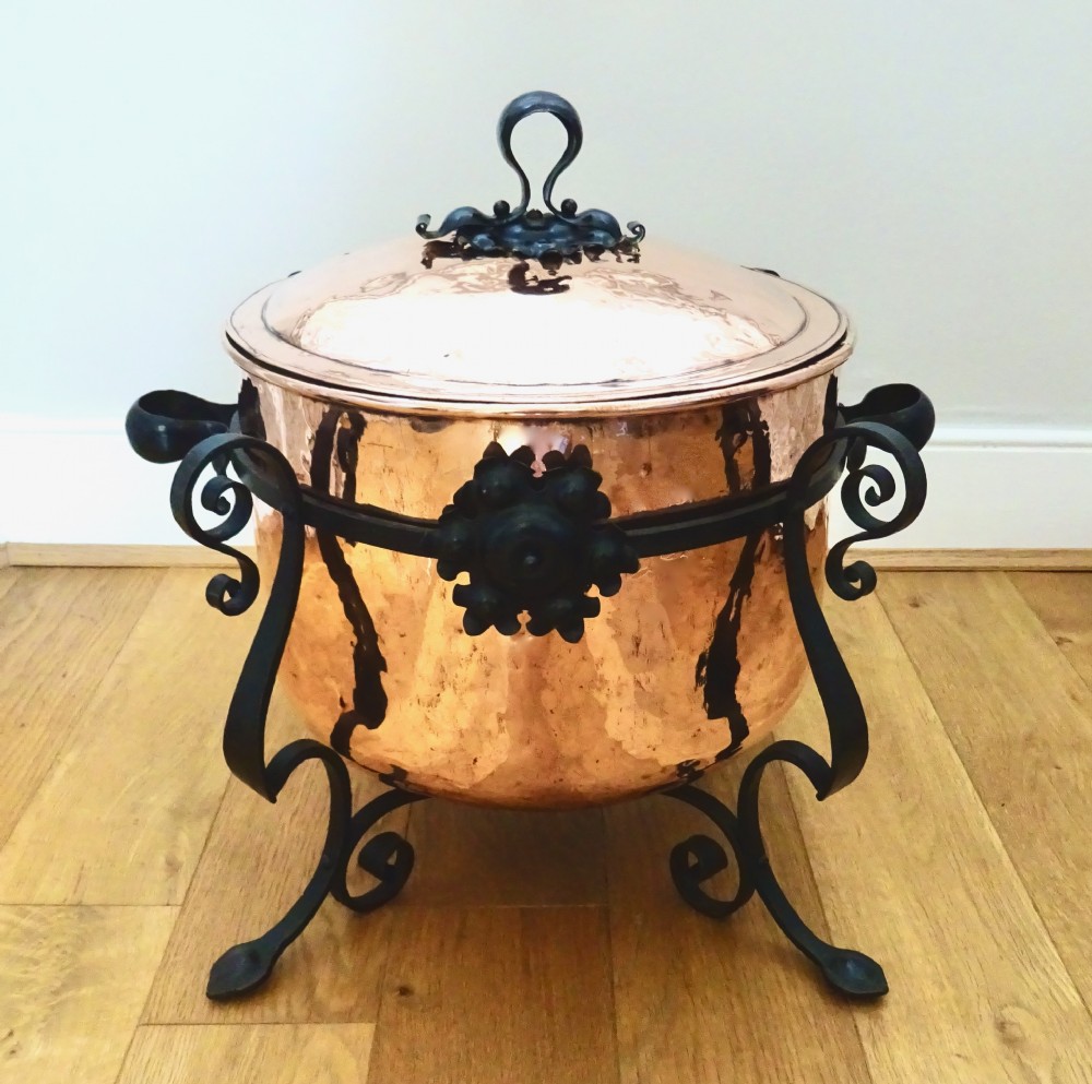 A large hammered copper bin with a domed lid fitted inside a black wrought iron stand with scrolled legs.