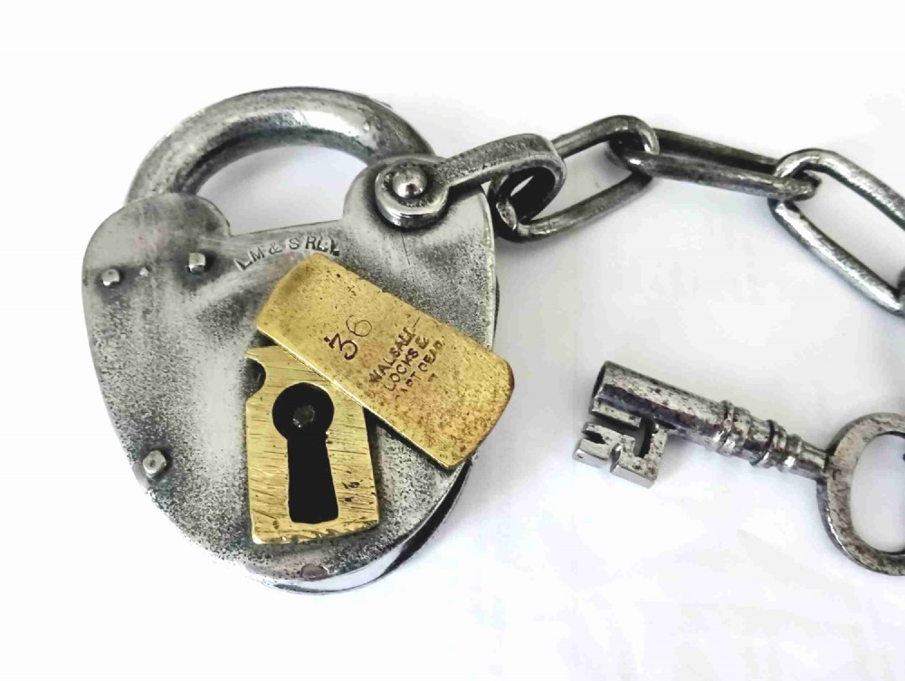 An antique heart-shaped steel railway padlock featuring a stamped brass keyhole cover, attached chain, and matching iron key.