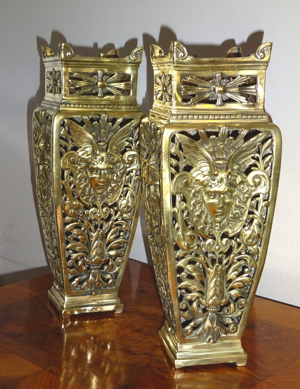 A pair of tall rectangular brass vases featuring pierced openwork scrolling foliage and central winged face motifs.