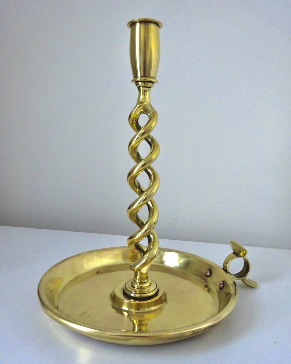 A polished brass chamberstick featuring an open barley twist stem set into a circular dish tray with a thumb ring handle.