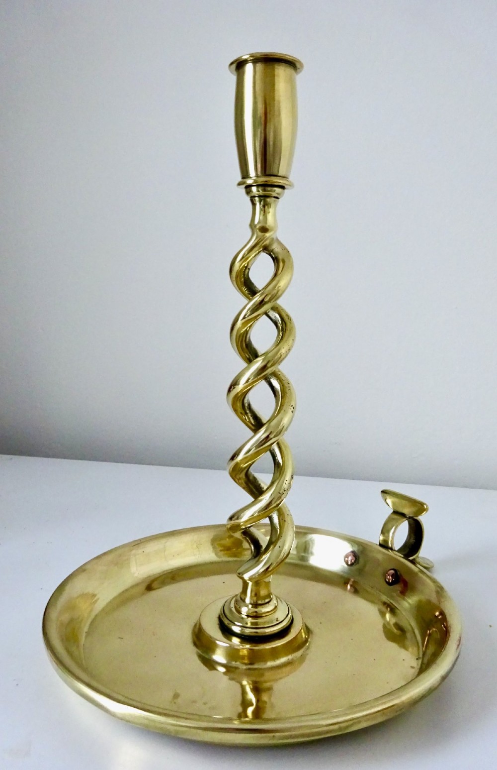 A polished brass chamberstick featuring an open barley twist stem set into a circular dish tray with a thumb ring handle. View VI