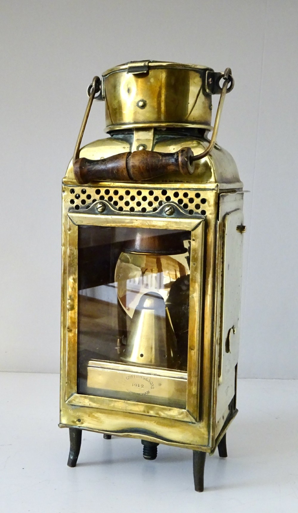 An antique brass oil lantern featuring a rectangular glass window, perforated ventilation top, turned wooden handle, and stamped manufacturer markings.