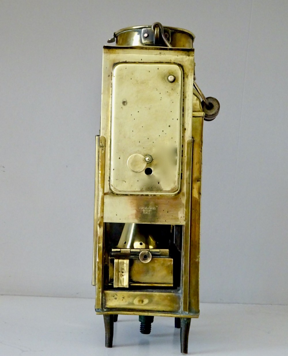 An antique brass oil lantern featuring a rectangular glass window, perforated ventilation top, turned wooden handle, and stamped manufacturer markings. View V