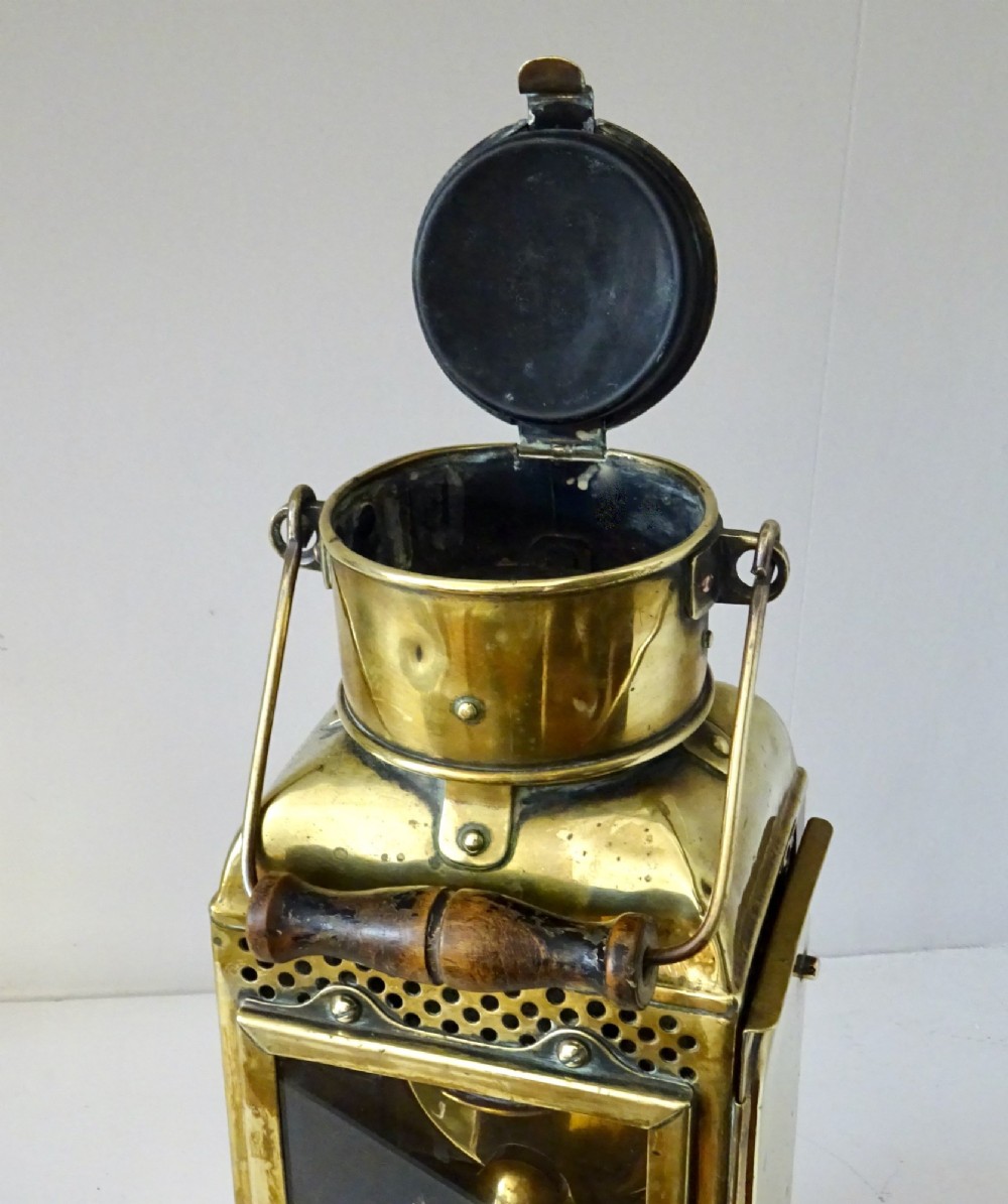 An antique brass oil lantern featuring a rectangular glass window, perforated ventilation top, turned wooden handle, and stamped manufacturer markings. View VI