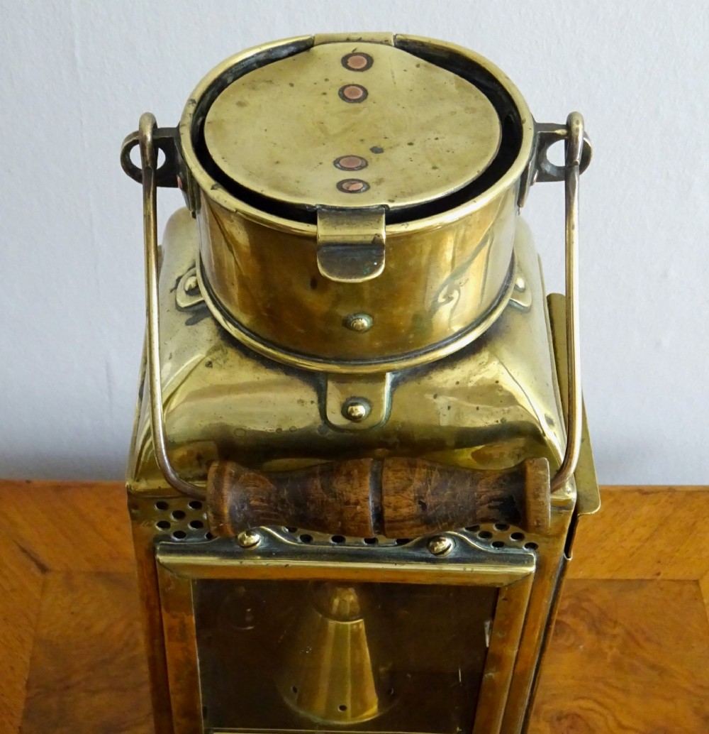 An antique brass oil lantern featuring a rectangular glass window, perforated ventilation top, turned wooden handle, and stamped manufacturer markings. View VII