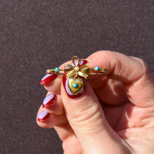 fig 2. An antique yellow gold bar brooch featuring a central bow motif with a seed pearl, a dangling gold heart set with a turquoise cabochon, and two flanking turquoise stones.