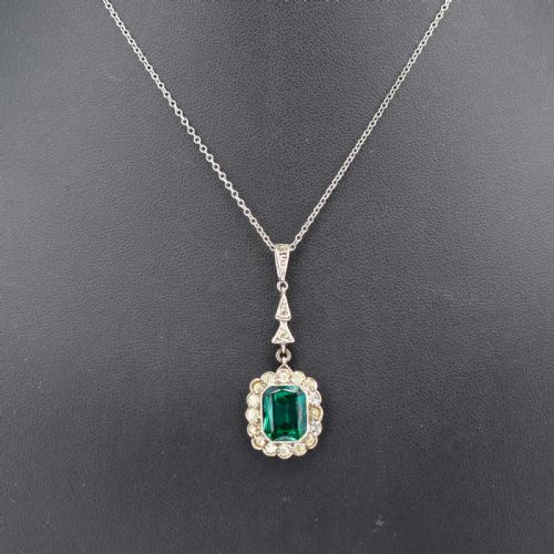 fig 1. An antique silver pendant necklace featuring an emerald-cut green paste stone surrounded by a clear paste stone halo, suspended from an articulated linear drop.