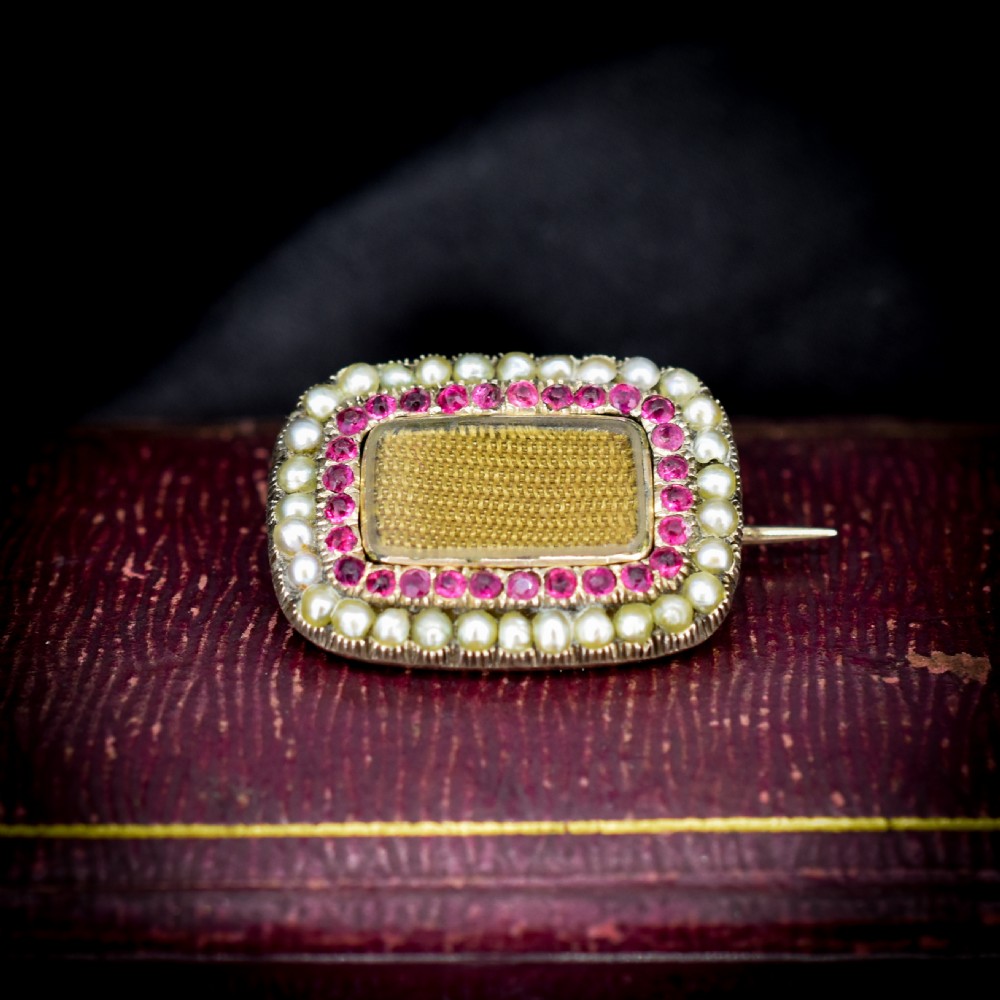 A rectangular Georgian gold mourning brooch featuring a central woven hair panel under glass, framed by a border of ruby paste stones and an outer rim of seed pearls.