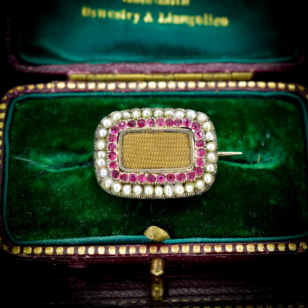 A rectangular Georgian gold mourning brooch featuring a central woven hair panel under glass, framed by a border of ruby paste stones and an outer rim of seed pearls. View II