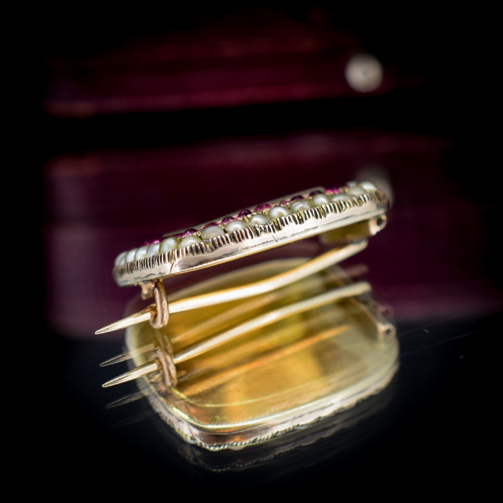 A rectangular Georgian gold mourning brooch featuring a central woven hair panel under glass, framed by a border of ruby paste stones and an outer rim of seed pearls. View V