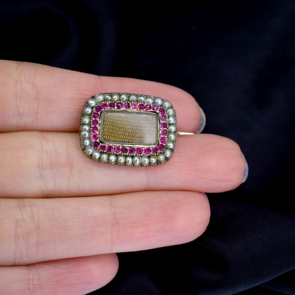 A rectangular Georgian gold mourning brooch featuring a central woven hair panel under glass, framed by a border of ruby paste stones and an outer rim of seed pearls. View VI