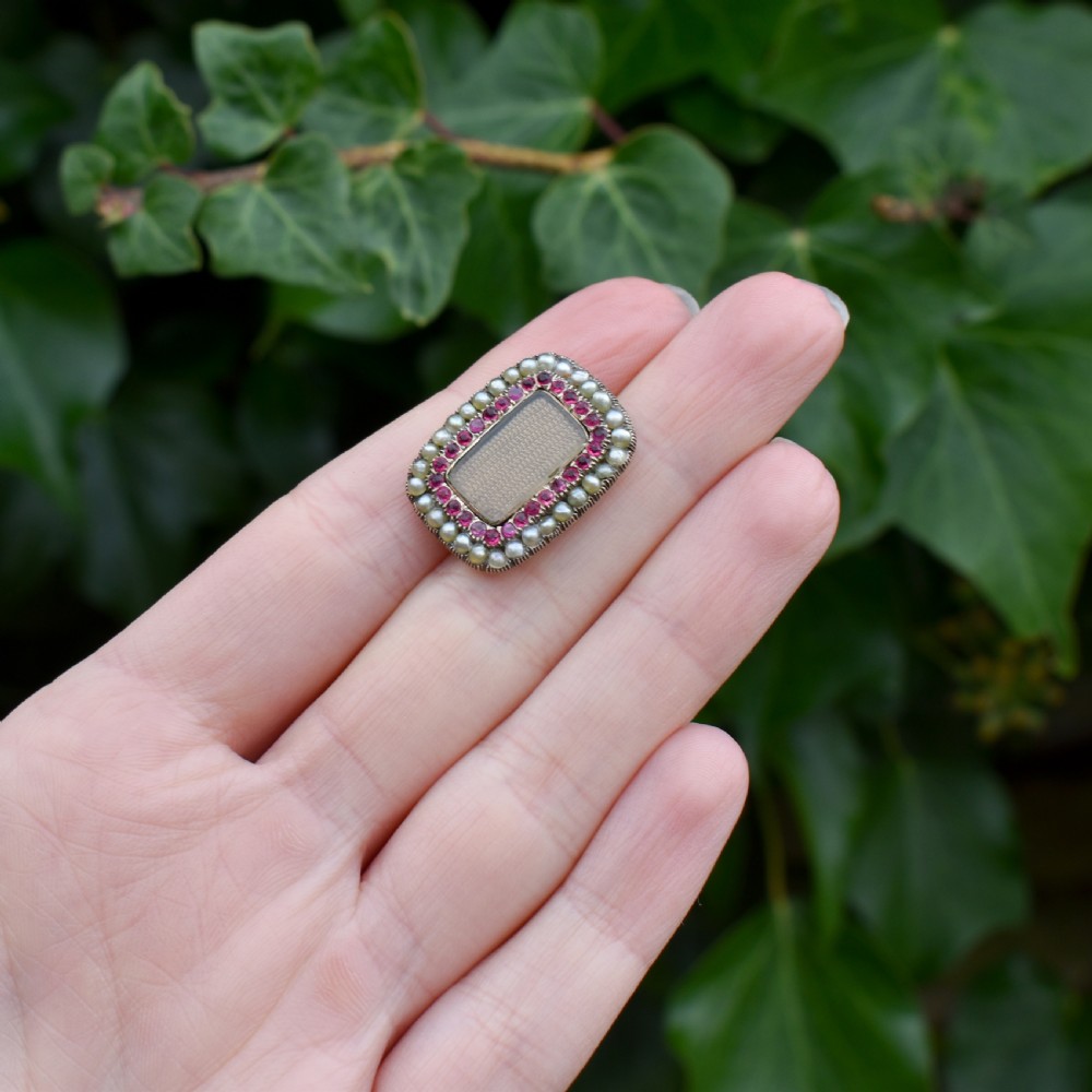 A rectangular Georgian gold mourning brooch featuring a central woven hair panel under glass, framed by a border of ruby paste stones and an outer rim of seed pearls. View X