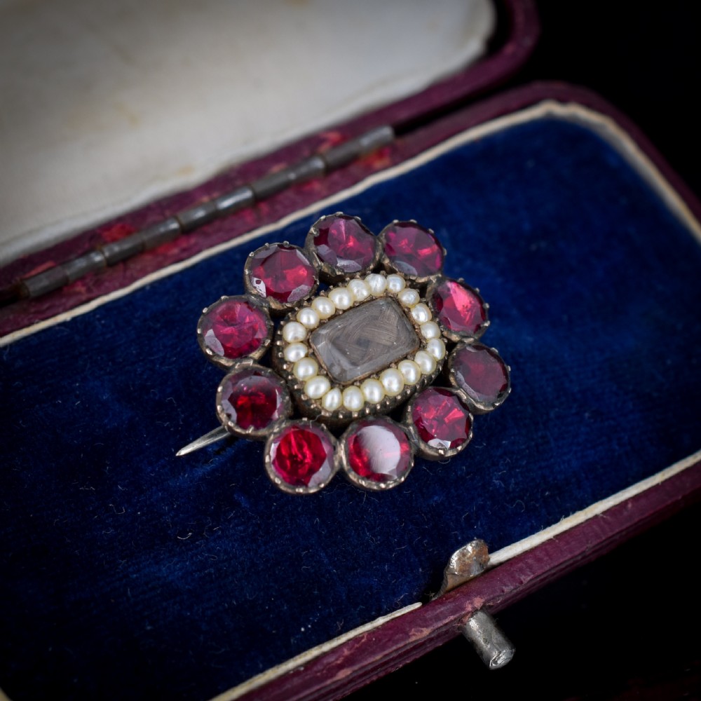 A Georgian cluster brooch featuring flat-cut red garnets surrounding a halo of seed pearls and a central hairwork compartment set in gold.