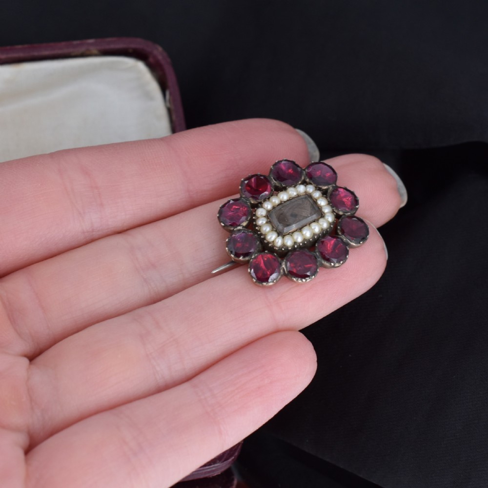 A Georgian cluster brooch featuring flat-cut red garnets surrounding a halo of seed pearls and a central hairwork compartment set in gold. View VIII