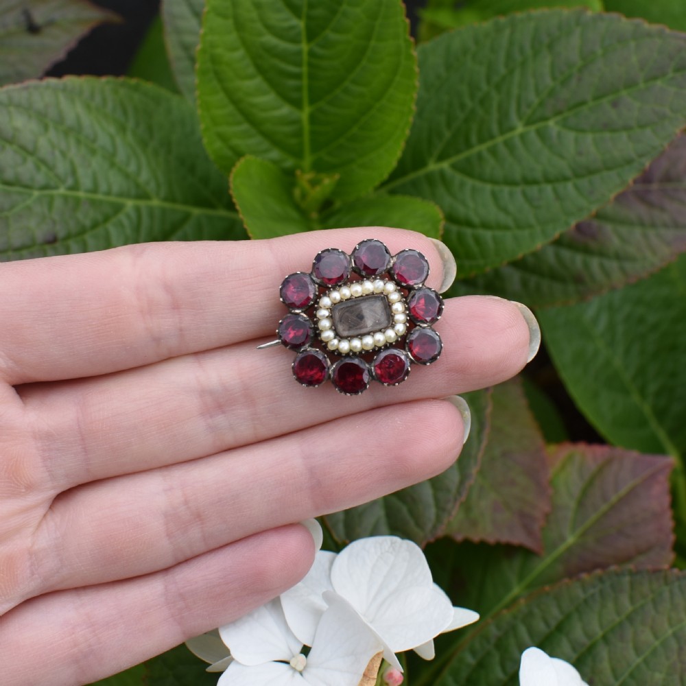 A Georgian cluster brooch featuring flat-cut red garnets surrounding a halo of seed pearls and a central hairwork compartment set in gold. View IX
