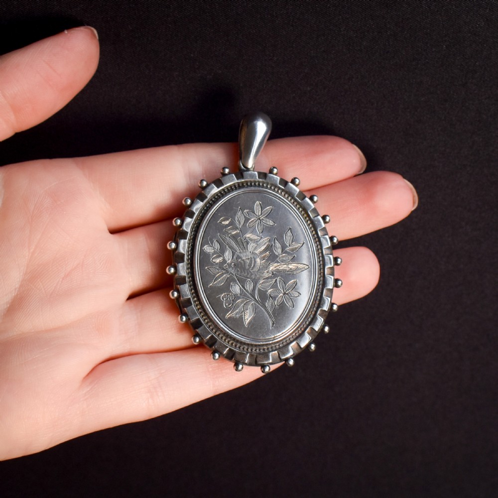 An oval sterling silver locket pendant featuring an engraved songbird amongst foliage surrounded by a beaded border. View II