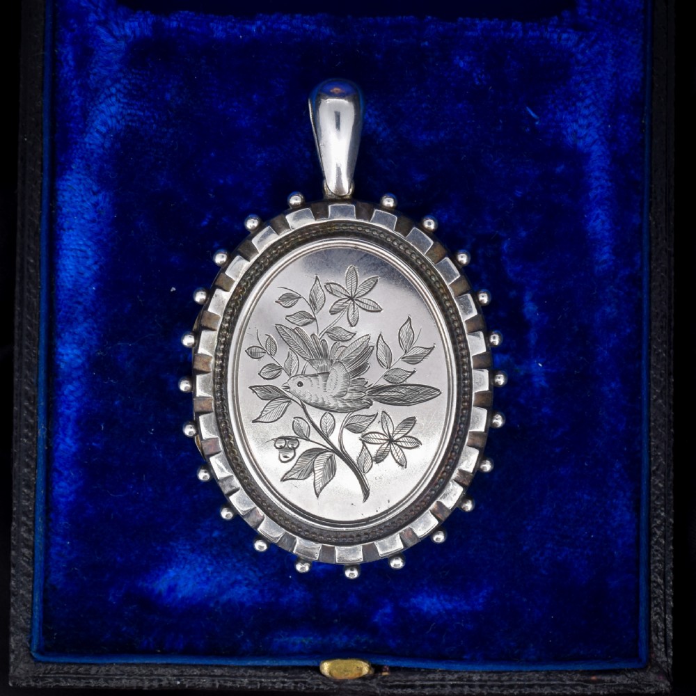 An oval sterling silver locket pendant featuring an engraved songbird amongst foliage surrounded by a beaded border.