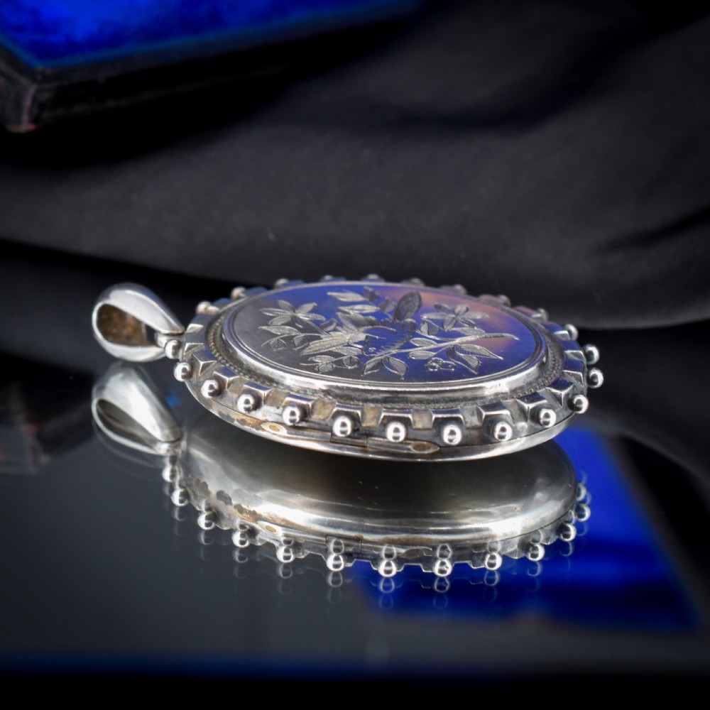 An oval sterling silver locket pendant featuring an engraved songbird amongst foliage surrounded by a beaded border. View V